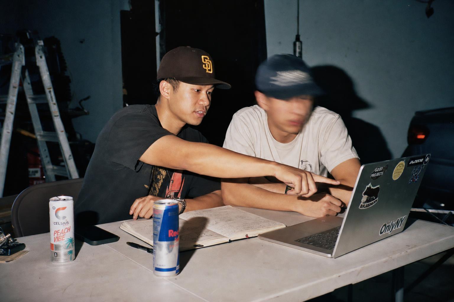 Two friends at a folding table in a dark studio, one explaining at a laptop