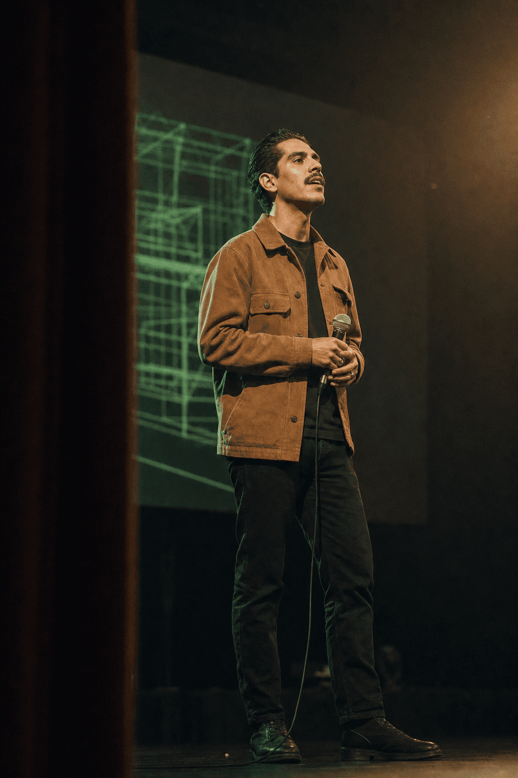 A host on a theater stage, an agent’s build projected huge behind him